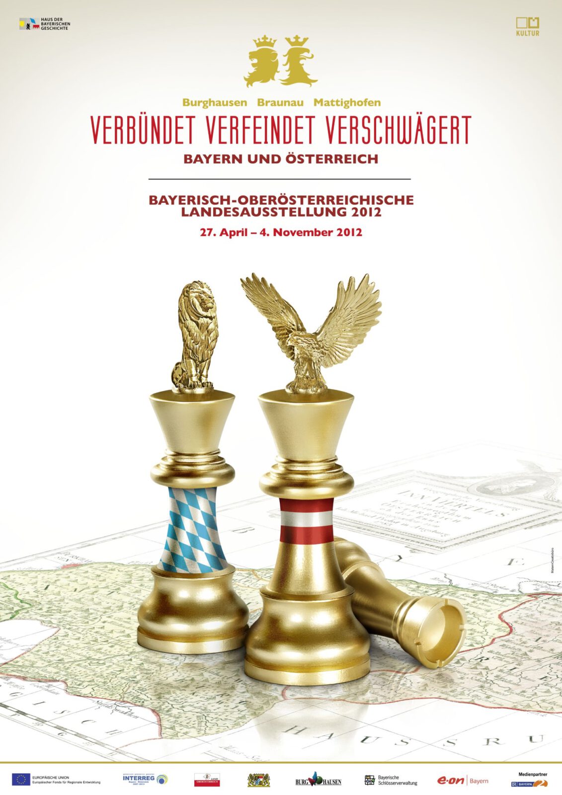 Logo der Bayerisch-Oberösterreichischen Landesausstellung 2012 in Burghausen, Braunau und Mattighofen. Drei Schachfiguren auf einer Landkarte. Eine stehende Schachfigur mit dem bayerischen Löwen und den weiß-blauen Rauten. Eine zweite stehende Schachfigur mit dem österreichischen Adler und rot-weiß-roten Streifen. Die dritte Schachfigur liegt .