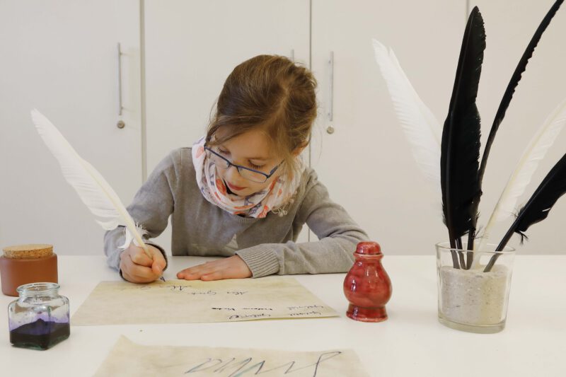 Ein Mädchen sitzt an einem Tisch. Sie schreibt mit einer Feder Buchstaben auf ein Blatt Papier.