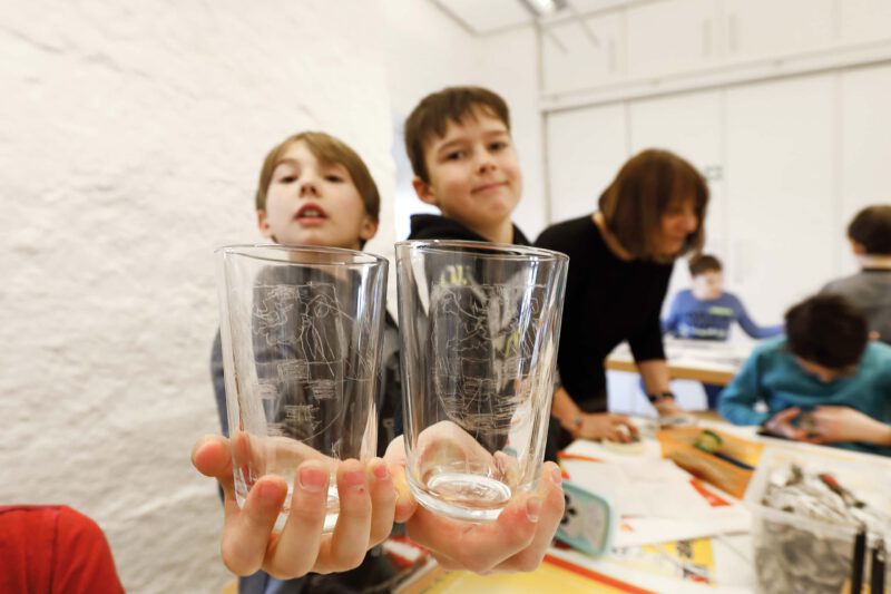 Zwei Jungen halten je ein Glas nach vorne. In die Gläser sind Wappen geritzt.