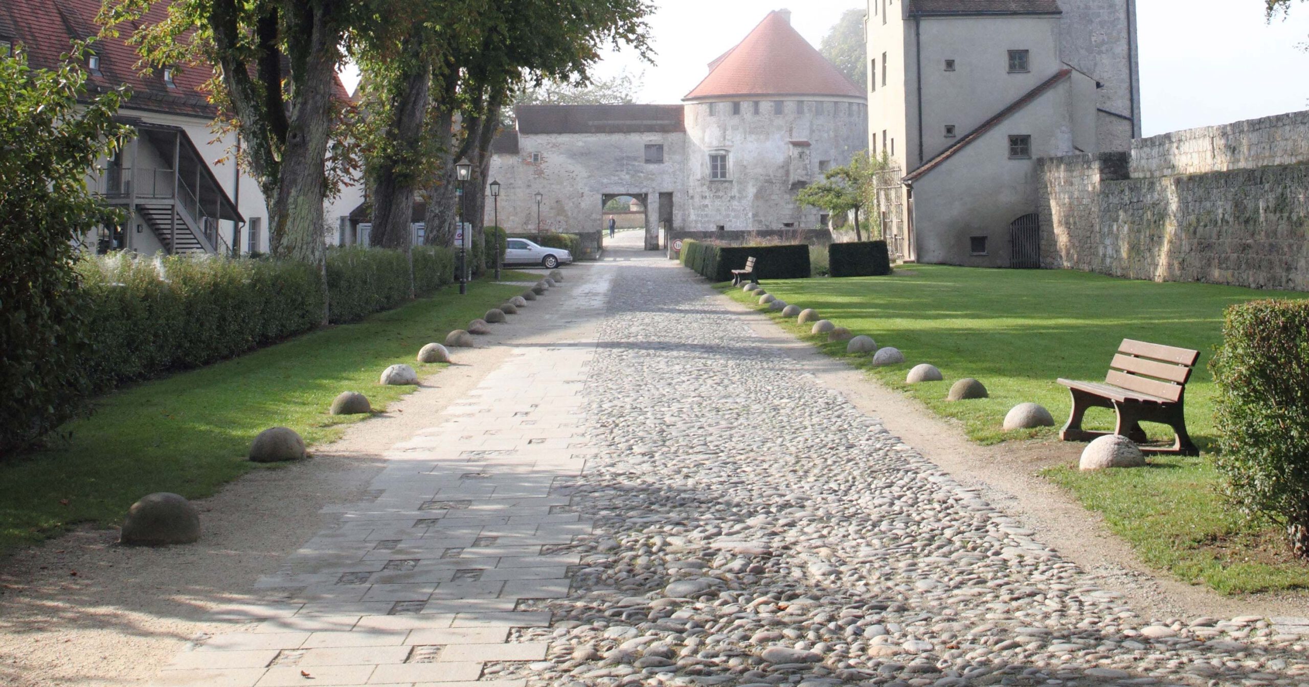 Barrierefreiheit Weg auf der Burganlage Der eine Teil des Wegs ist gepflastert, Der andere Teil ist mit großen Steinen gefüllt Im Hintergrund ist ein Burgtor zu sehen