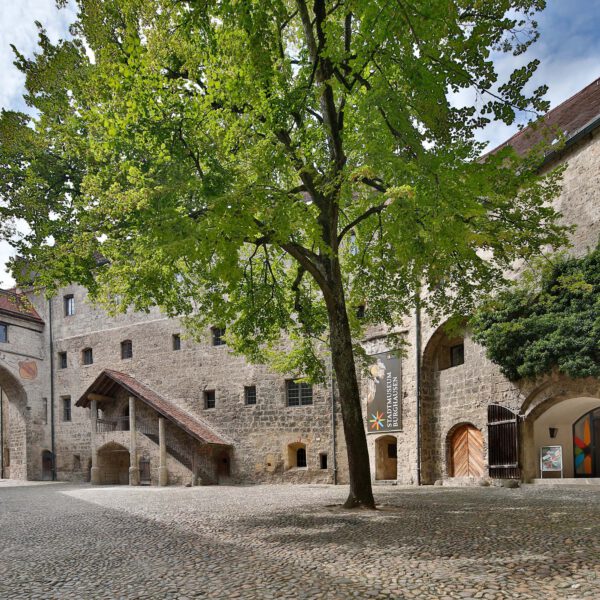 Stadtmuseum Burghausen Burghof Innenhof einer Burg aus Stein In der Mitte des Hofs steht ein Baum Rechts ist ein Eingang mit einem Plakat Stadtmuseum Burghausen