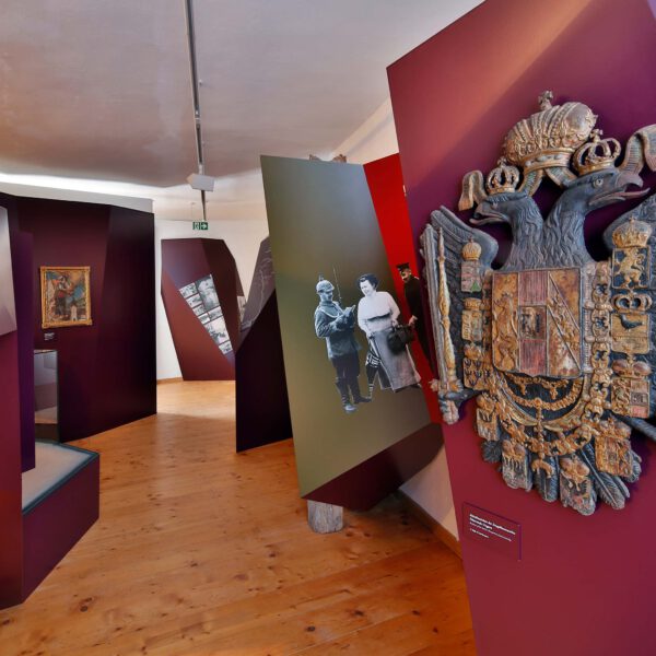 Ausstellung Stadtgeschichte mit roten Stellwänden in unterschiedlichen Formen. Das Wappen von Österreich mit dem Doppeladler aus Eisen hängt an einer Wand.