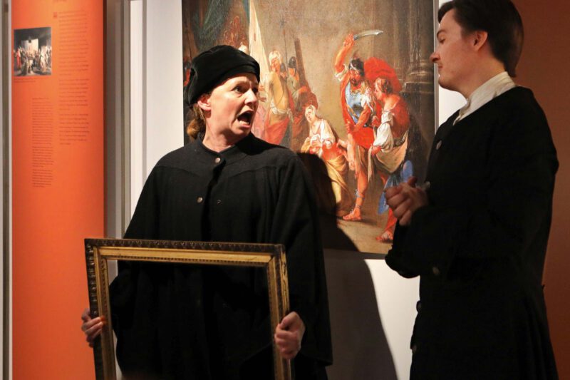 Vor einem Gemälde stehen zwei schwarz gekleidete Personen. Eine hält einen Bilderrahmen in der Hand.